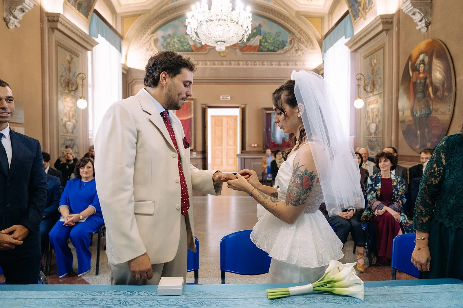 A couple exchanging wedding rings during a wedding ceremony in an ornate church, with guests seated in the background.