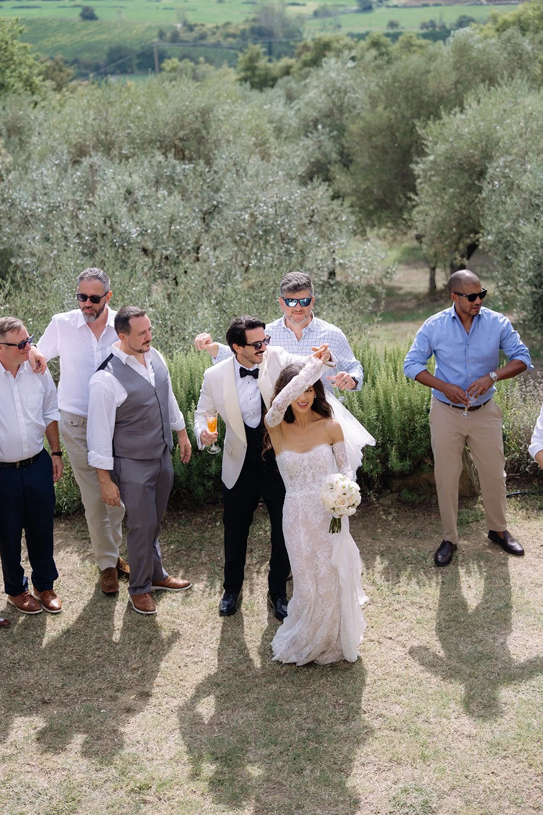 A bride and groom celebrating outdoors with friends in a vineyard, some people wearing sunglasses, holding drinks, and smiling.