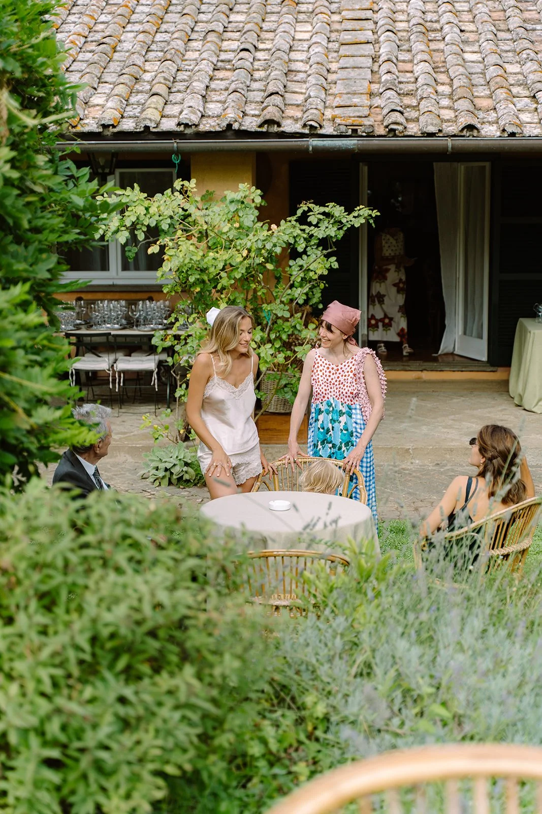 A group of people, including two women standing and talking, sitting around a round outdoor table in a garden or backyard, with lush greenery and a building with a tiled roof in the background.