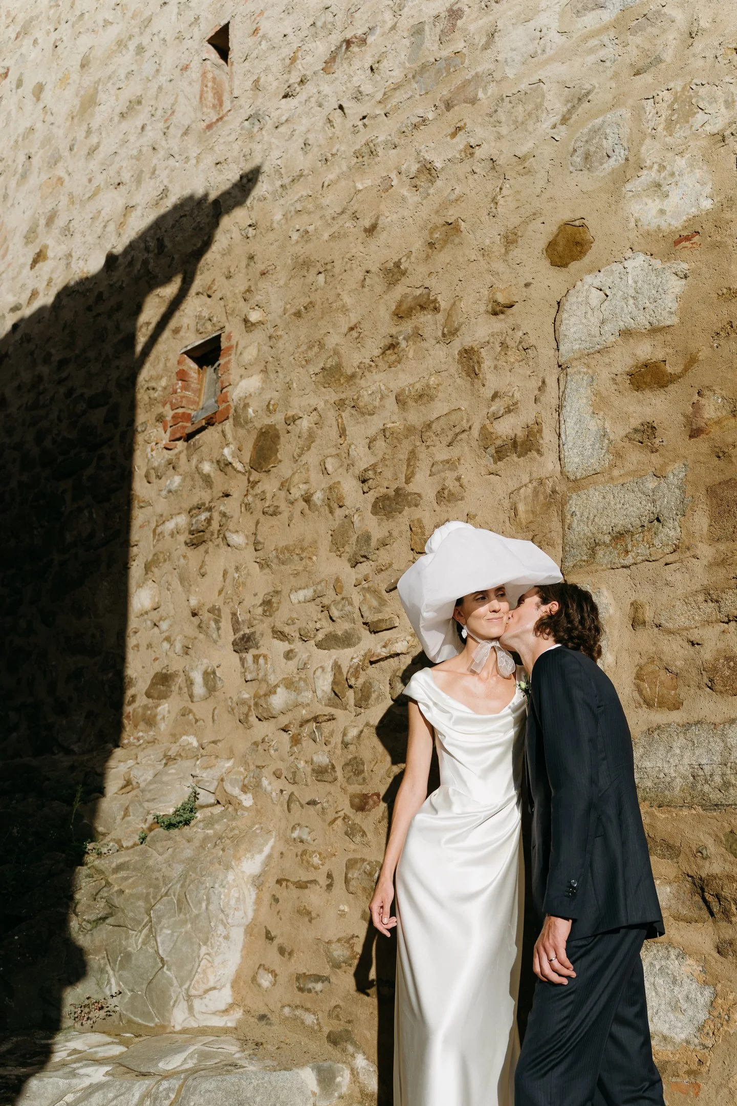 A couple dressed in wedding attire sharing a kiss, the woman wearing a white dress and an oversized white hat, and the man in a black suit, standing against a rustic stone wall.
