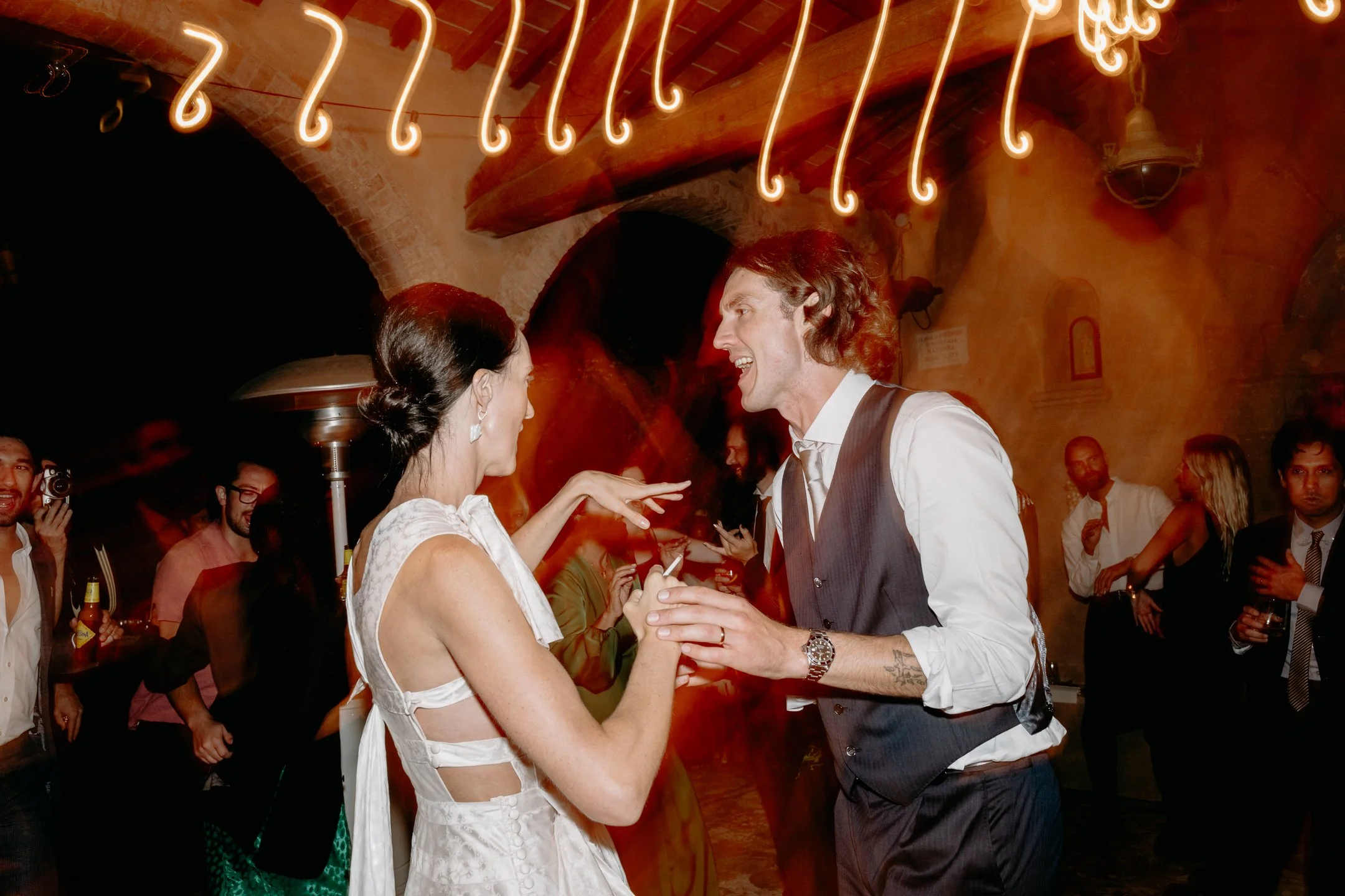 Two people dancing and laughing at a party, with other guests in the background under warm lighting.