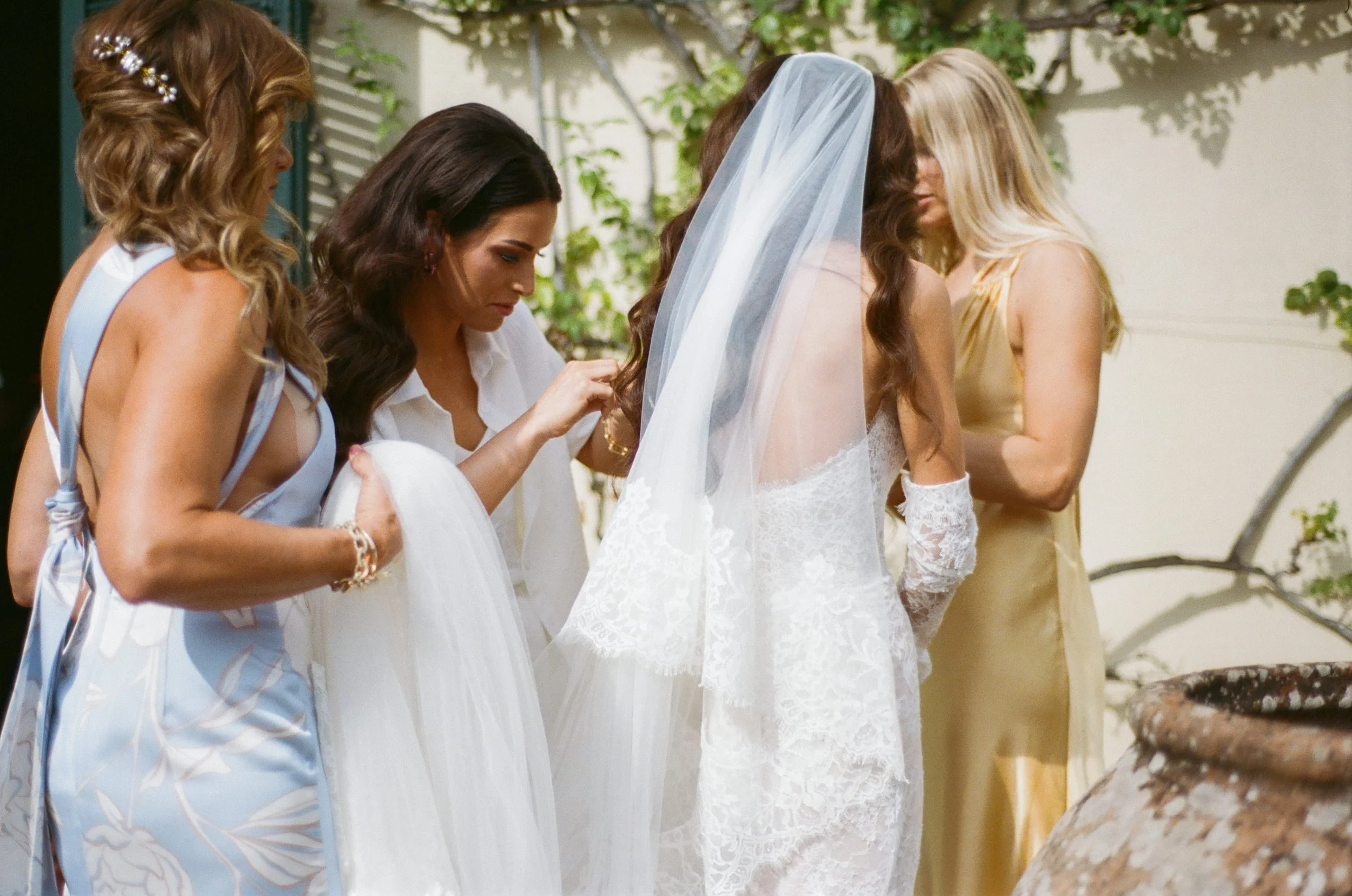 A bride getting ready for her wedding with her bridesmaids outdoors, one bridesmaid adjusting the bride's dress, all in elegant dresses.
