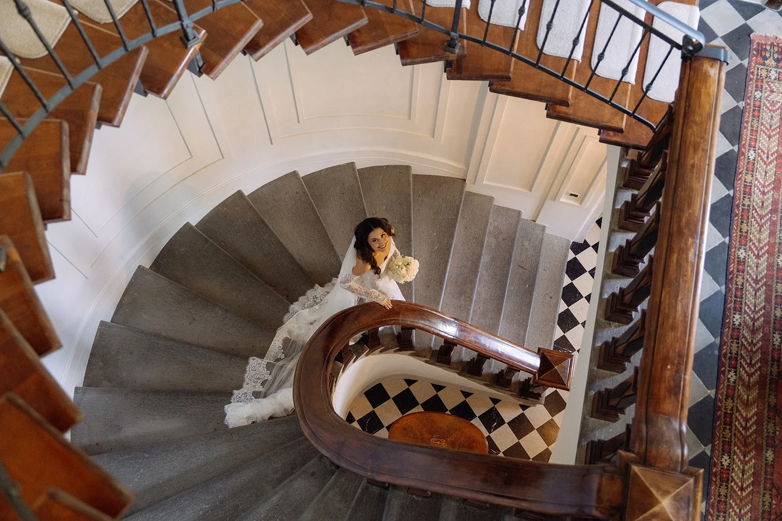 A bride in a white wedding dress holding a bouquet standing at the top of a wooden curved staircase inside a building.
