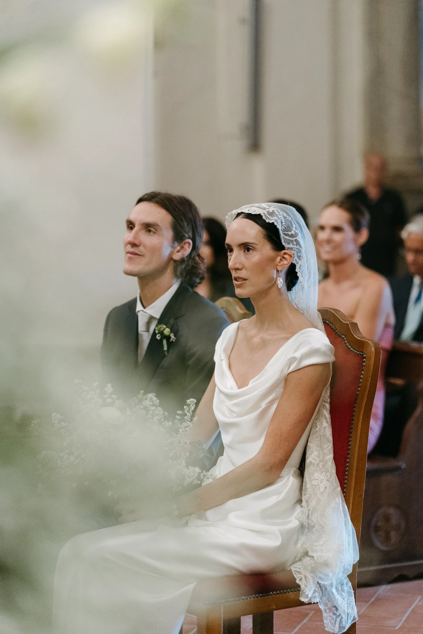 A bride and groom sitting during a wedding ceremony inside a church, with several guests in the background.