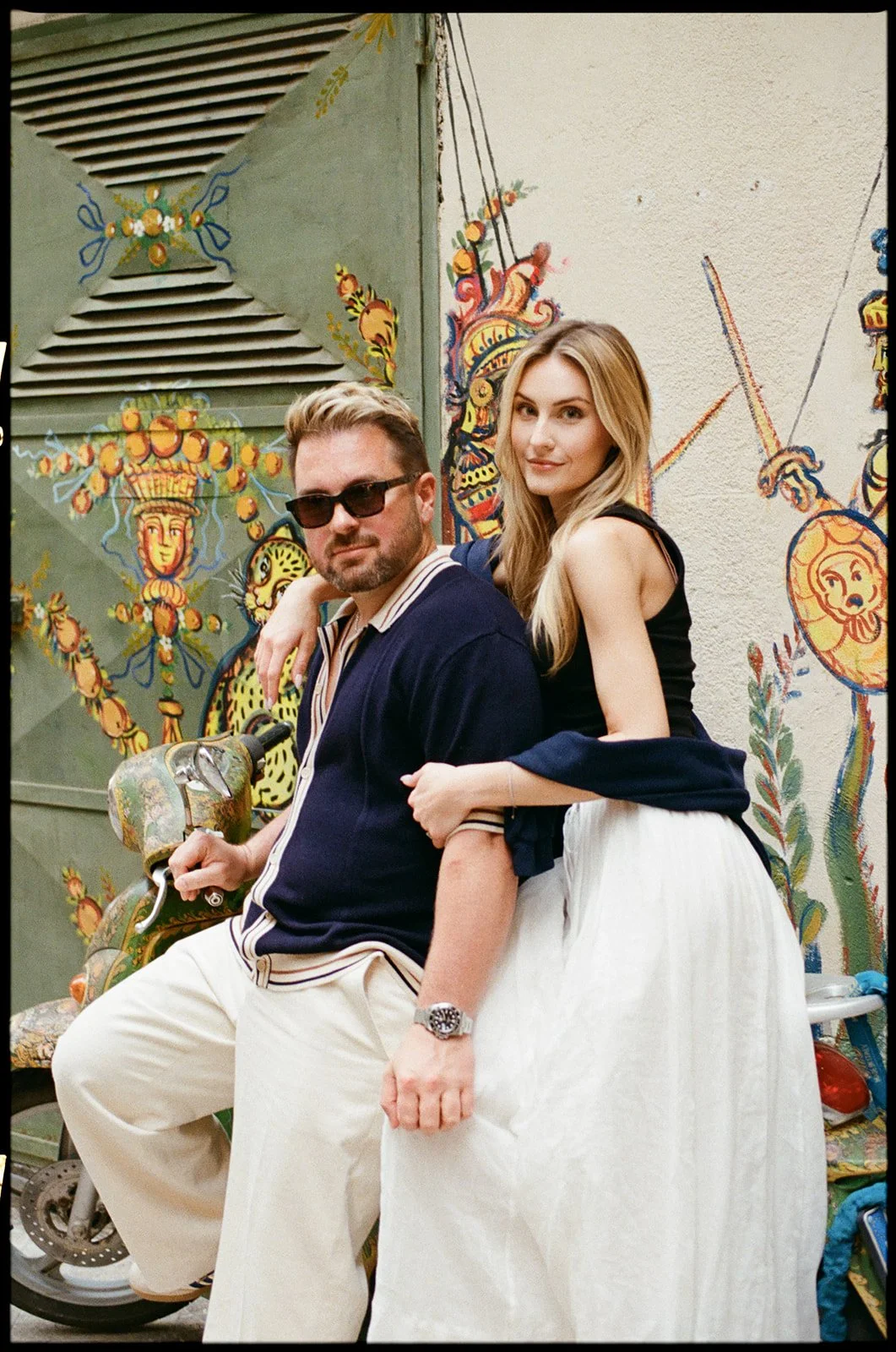 A man with sunglasses and a woman with long blonde hair posing together outdoors in front of a colorful, mural-style wall. The man is holding a vintage-style scooter and wears a navy jacket with a striped shirt underneath. The woman is leaning on his