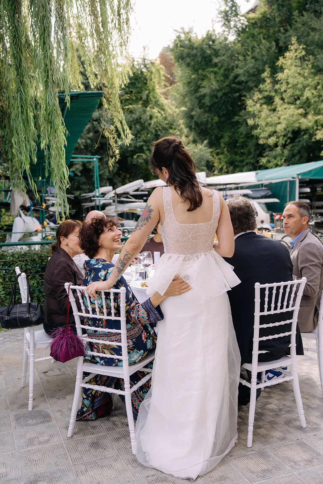 A woman in a white dress, likely a bride, standing and leaning toward seated guests, smiling and engaging with a woman in a colorful dress. The group is outdoors at a formal event, with trees and parked boats in the background.