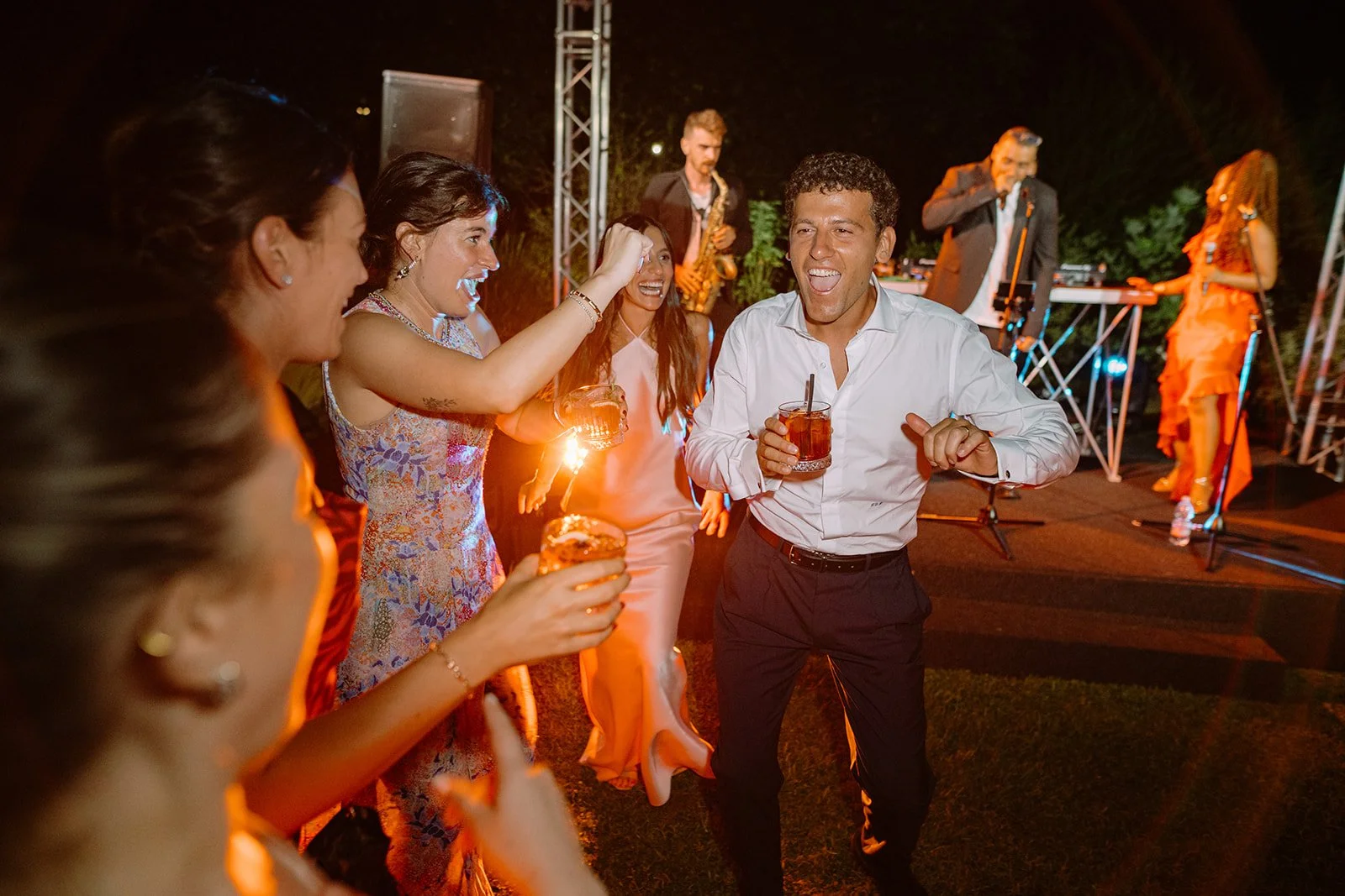 People celebrating at an outdoor party with a band playing in the background, dancing, and holding drinks during the night.