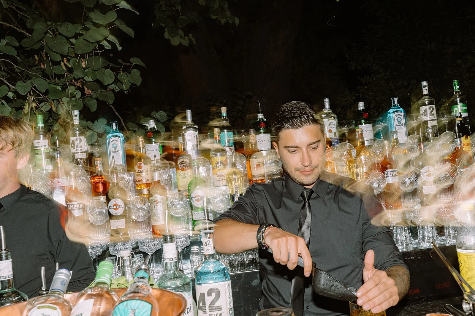 A bartender pouring a drink at an outdoor bar with numerous bottles of liquor and glasses behind him, illuminated at night.