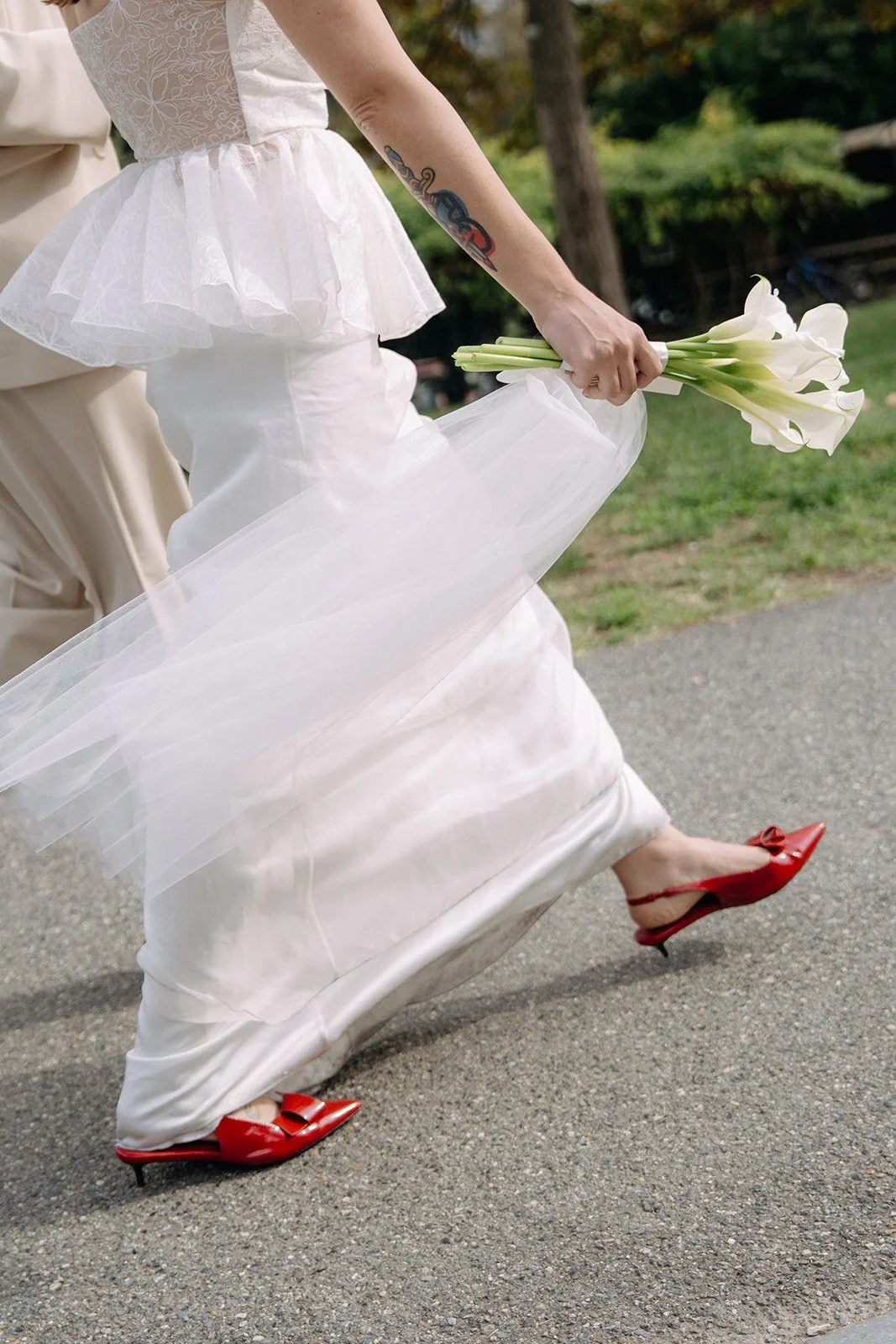 Close-up of a bride in a white wedding dress, holding a bouquet of white calla lilies, while lifting her dress to reveal red high heel shoes with bow details, walking on an outdoor path with green trees in the background.