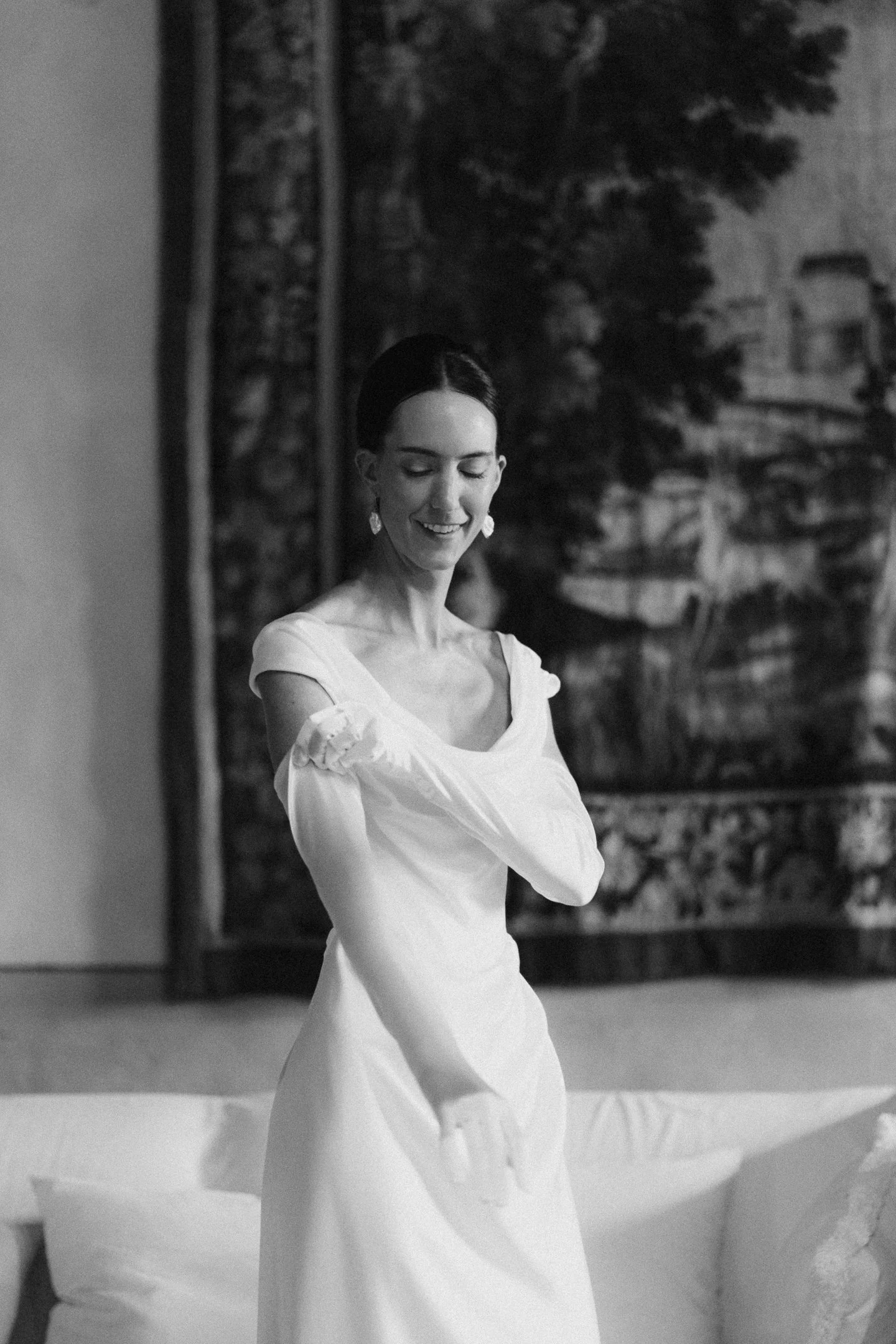 A smiling woman in a white dress with off-shoulder sleeves, standing indoors with a mural of buildings and trees in the background.