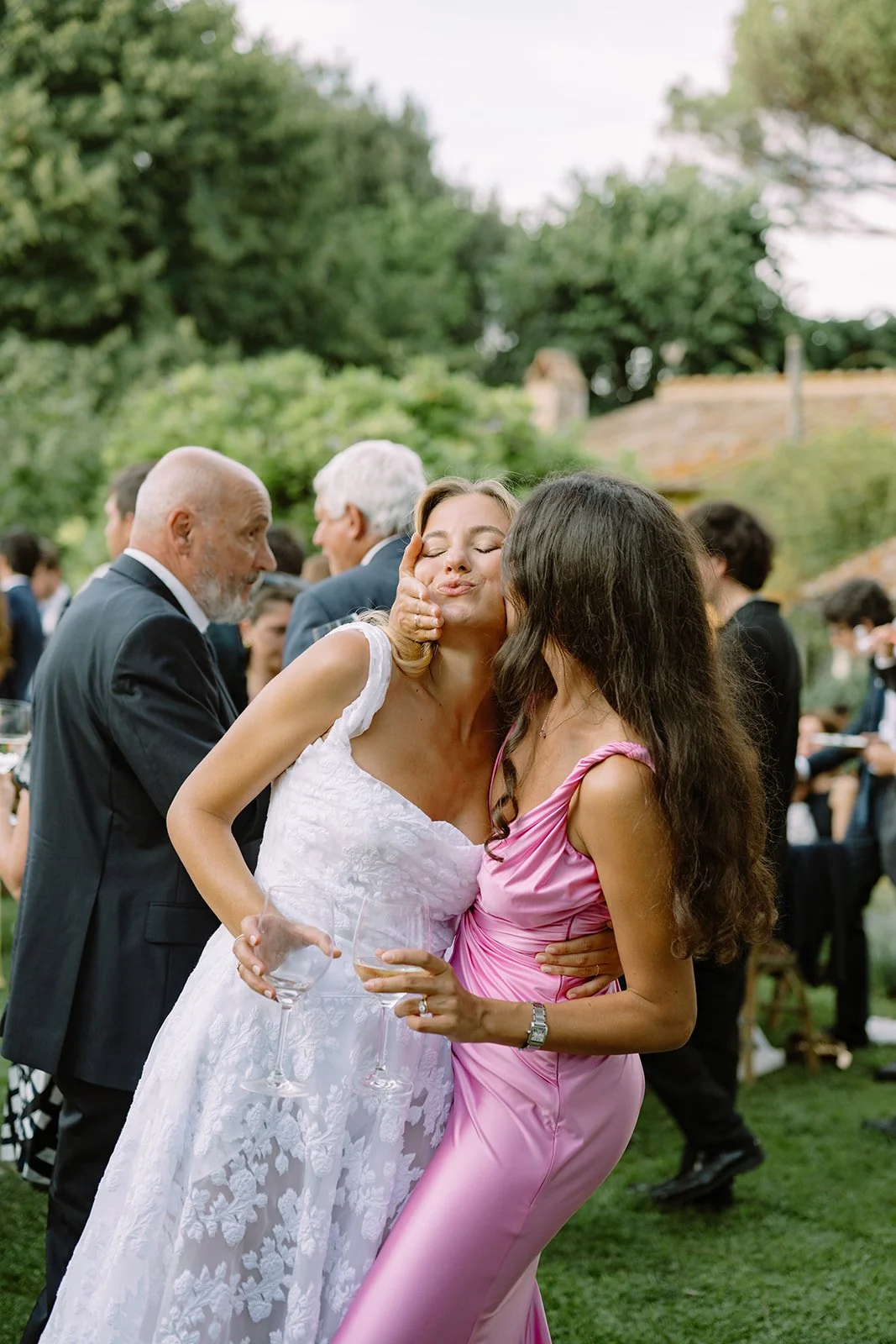 Two women, one in a white dress and the other in a pink dress, are hugging and sharing a kiss at an outdoor wedding reception.