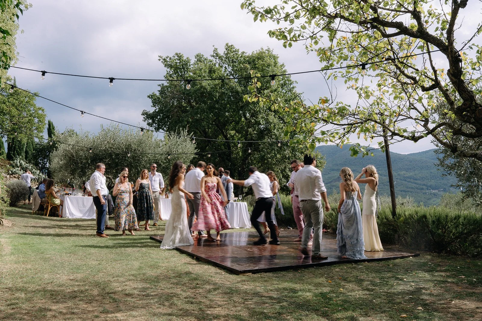 People dancing on a wooden dance floor at an outdoor event under string lights, with tables and a scenic view of greenery and mountains in the background.