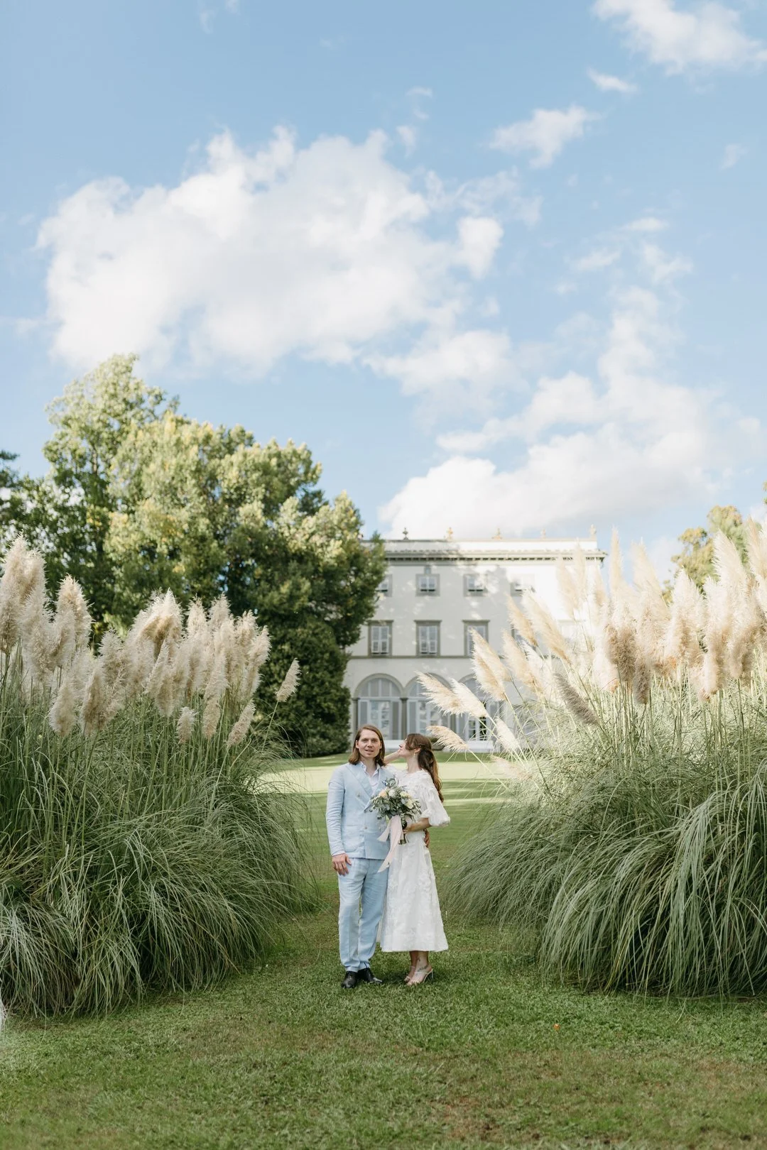 A man and woman dressed in wedding attire standing outdoors amidst tall grass and white pampas, with a large white building in the background and a blue sky with clouds above.