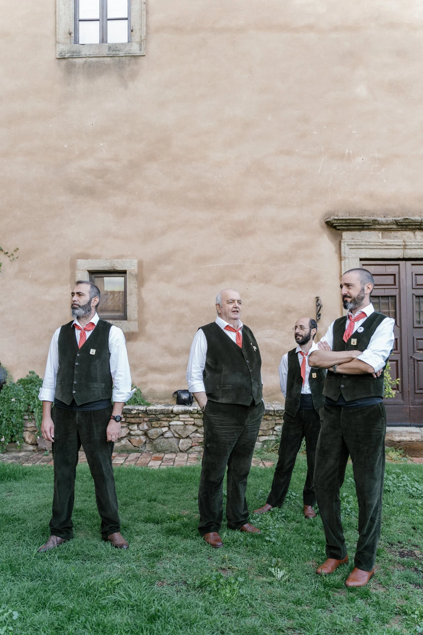 Four men dressed in formal attire with black vests, white shirts, red ties, and brown shoes, standing on a grassy lawn in front of an old stone building with small windows and a large wooden door.