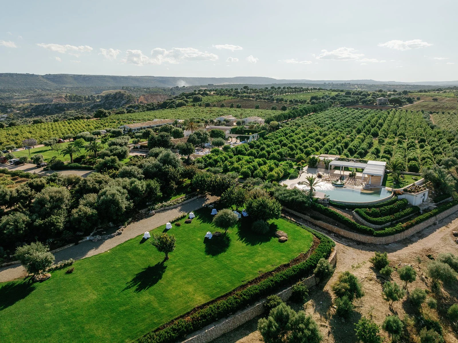 Aerial view of a lush, green estate with gardens, trees, a pool area, and surrounding agricultural fields under a partly cloudy sky.