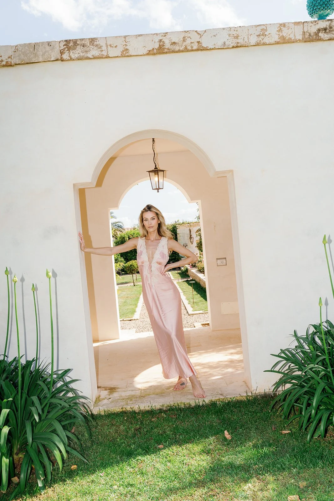 A woman in a pink satin dress standing in an arched doorway with greenery and a garden visible outside.