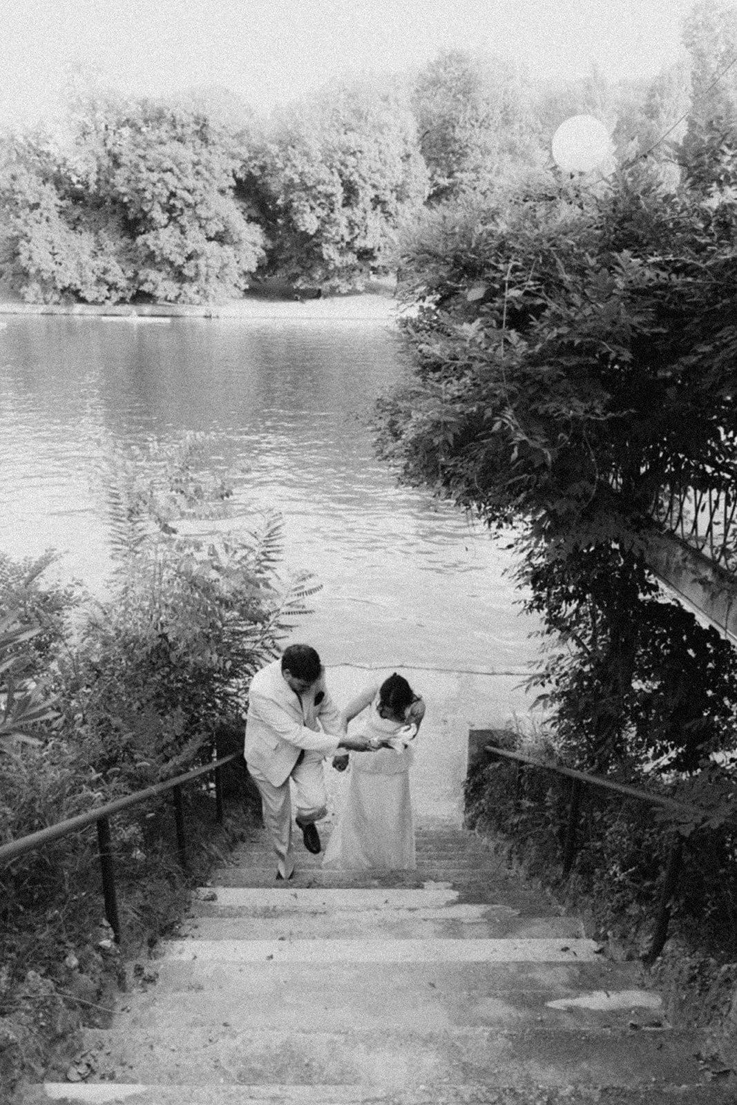 A man helping a woman up concrete stairs outside near a body of water, surrounded by trees and bushes, in black and white.