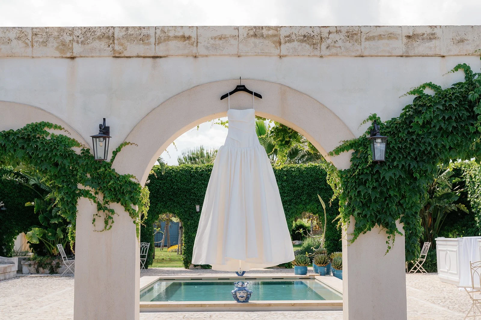 A white wedding dress hanging on a black hanger in front of an arched stone wall, against a background of greenery and a swimming pool.