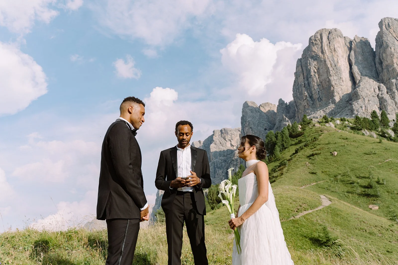 A wedding ceremony outdoors with a bride in a white dress holding calla lilies, and two men in suits, at the base of a mountain with green grass and rocky cliffs under a blue sky with white clouds.