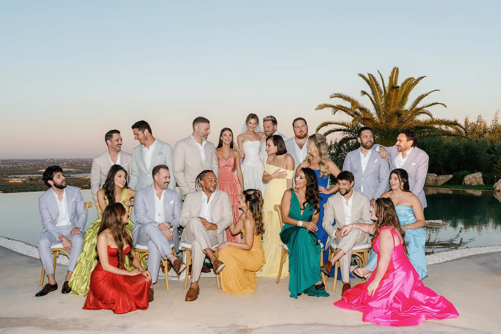 Group of people in colorful dresses and suits gathered outdoors near a pool with a palm tree in the background during sunset.