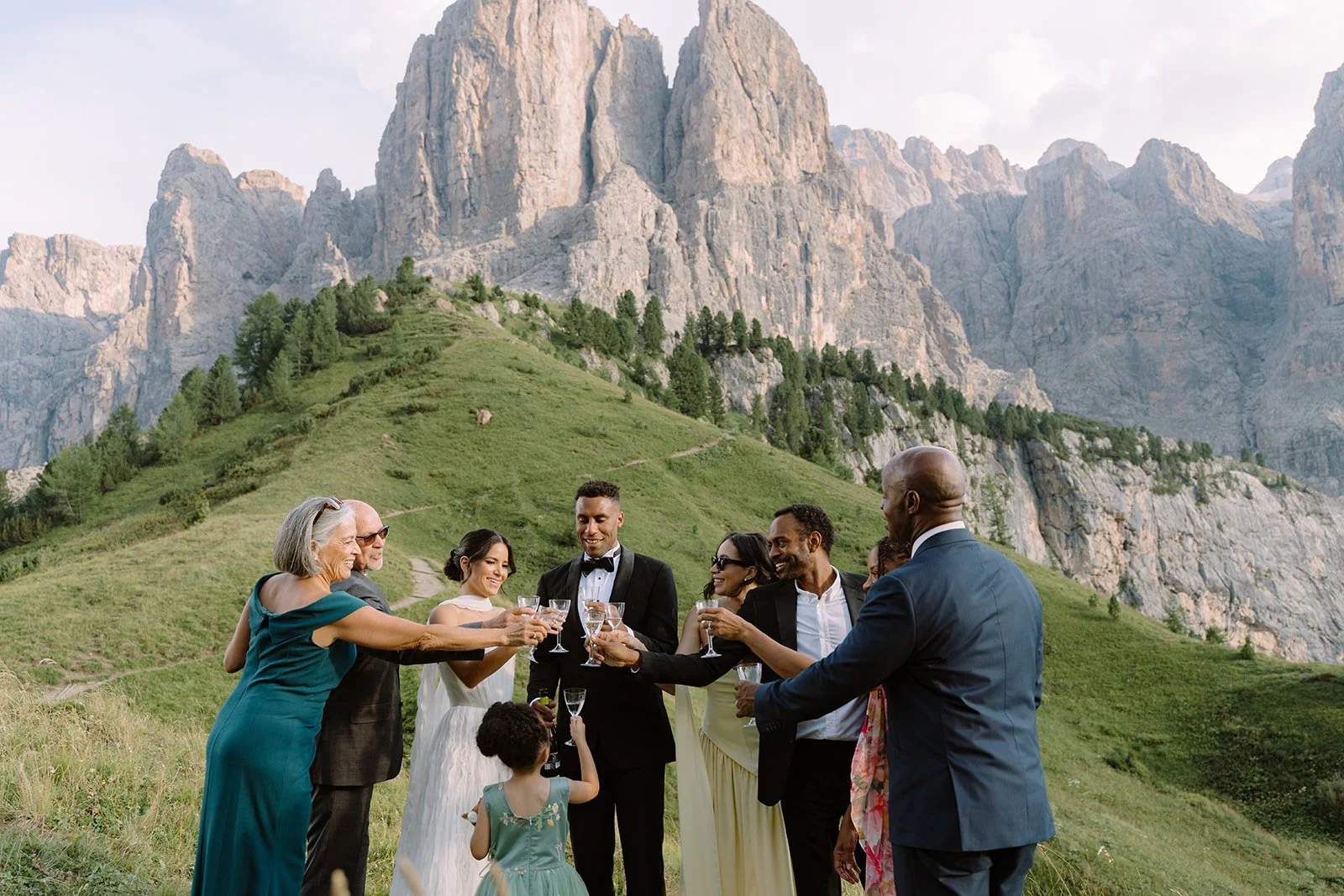 A group of people dressed in formal attire raising glasses for a toast outdoors, with mountains and greenery in the background.