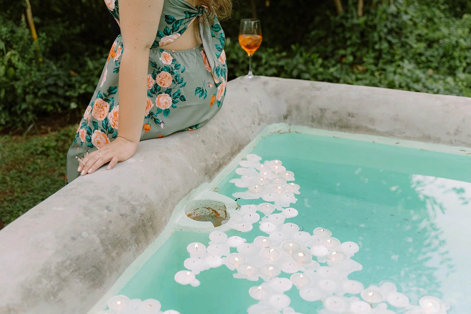A person leaning on a stone edge overlooking a water-filled fountain with floating white candles. The person is wearing a floral dress and has a glass of orange-colored drink nearby.