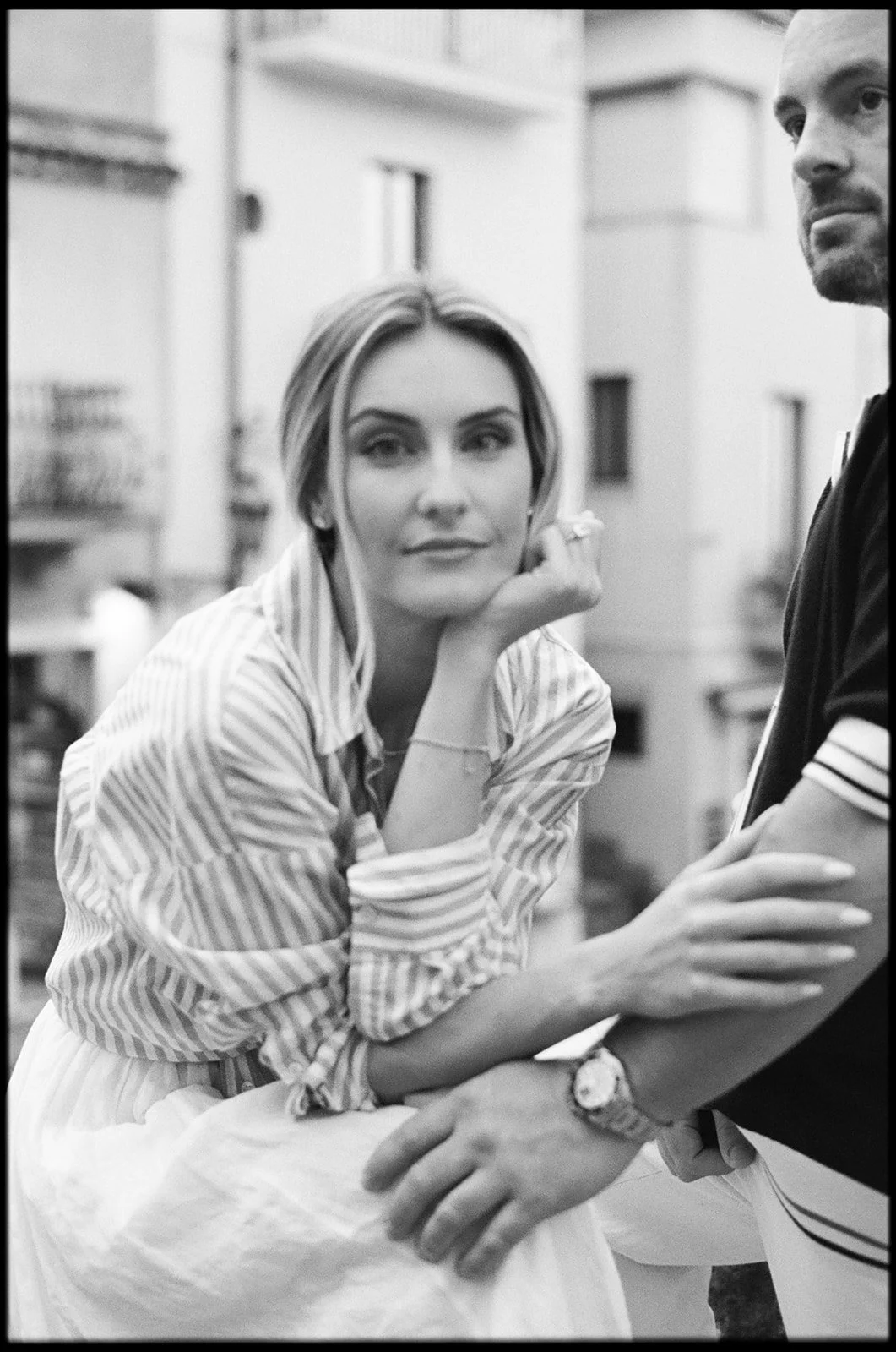A black-and-white photo of a woman with light-colored hair, resting her chin on her hand, looking thoughtfully at the camera. She is seated outdoors, with her arm supported by a man standing next to her, whose face is partially visible.