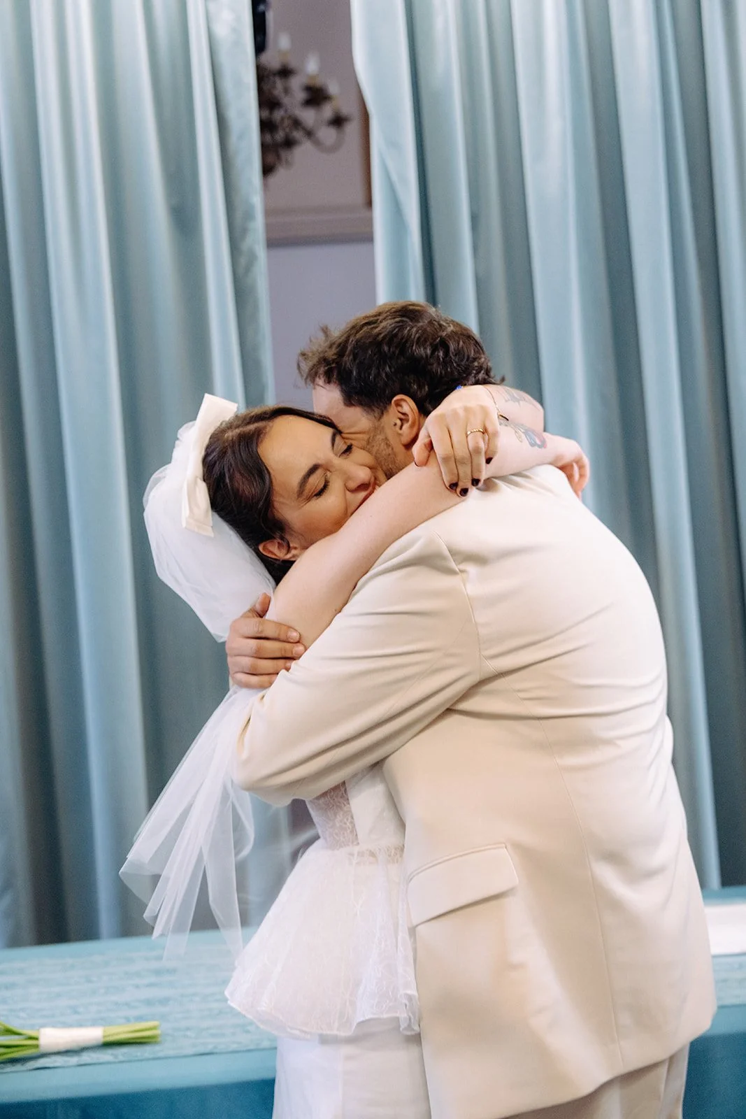 A woman in a wedding dress hugging a man in a beige suit during a wedding ceremony, with blue curtains in the background.