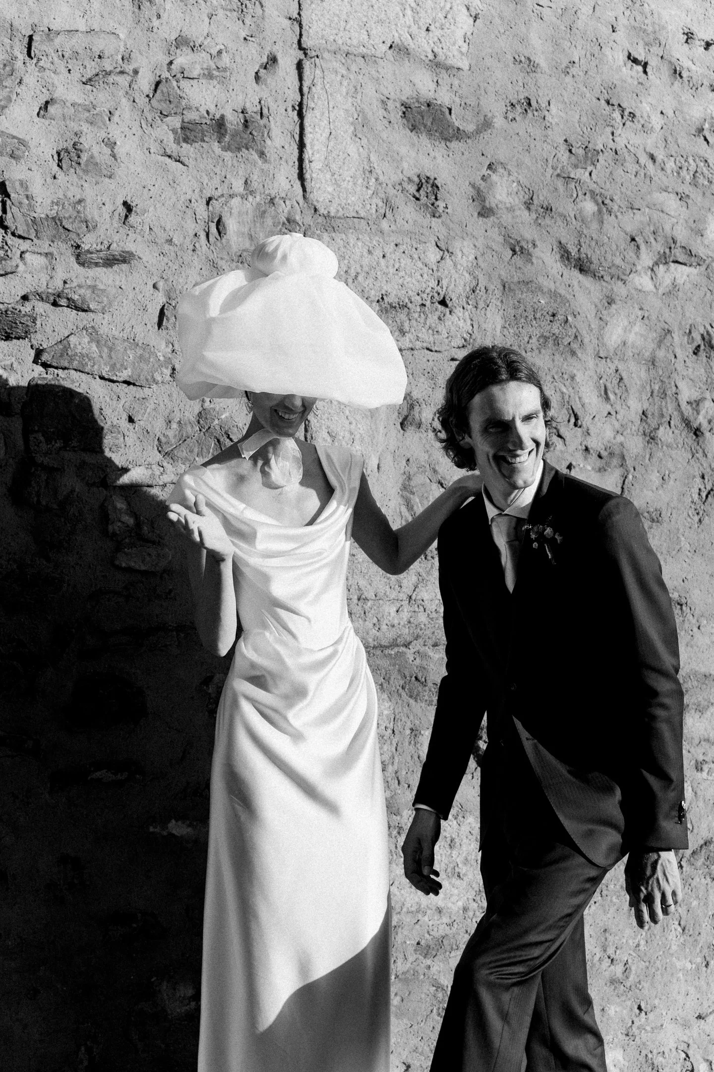 A bride and groom sharing a joyful moment, standing against a stone wall. The bride is wearing a white dress and a large fabric headpiece, and the groom is in a dark suit with a boutonniere.