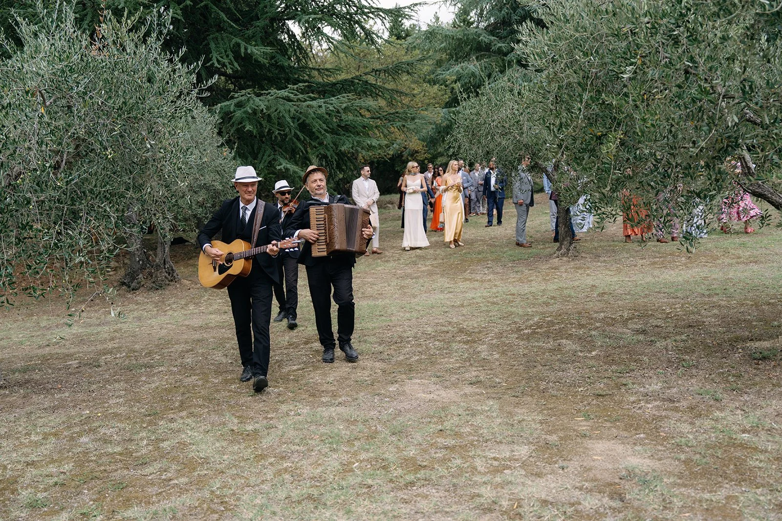 Musicians playing guitar and accordion walking through a garden with wedding guests in the background.