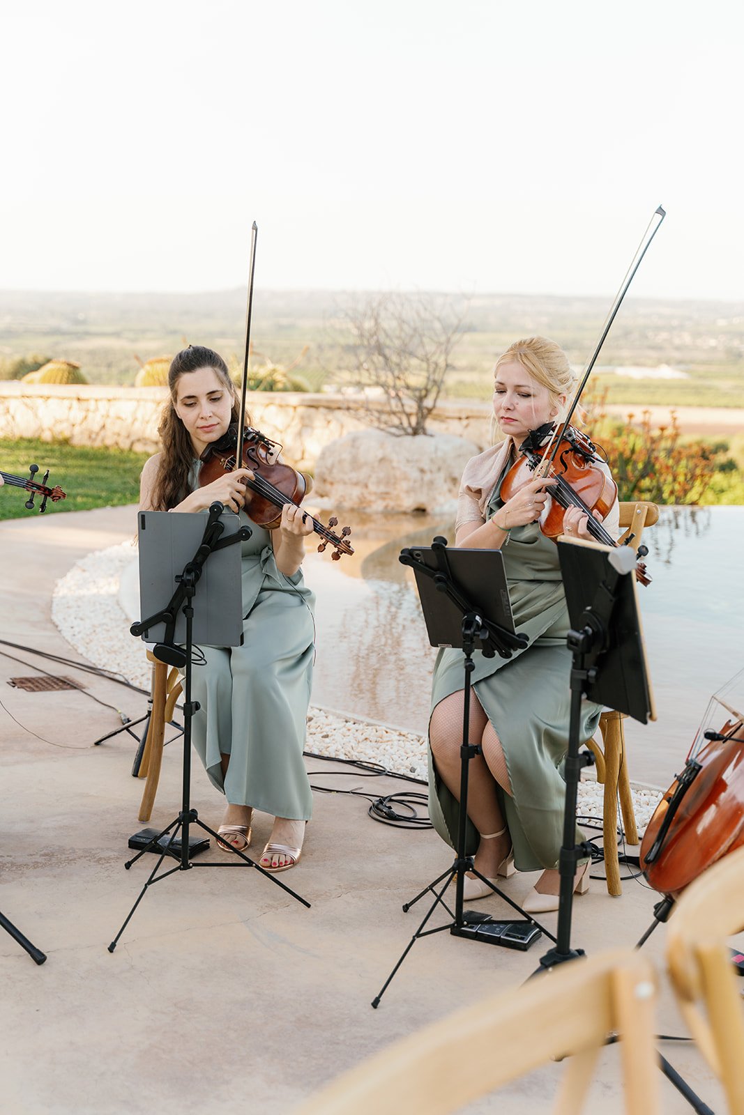 Two women playing violins outdoors during a musical performance, with music stands and a scenic landscape in the background.