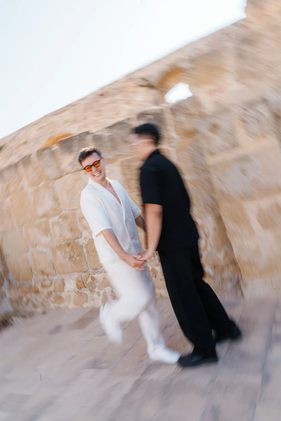 Two women holding hands, smiling and laughing, standing outdoors against a stone wall with an angled background, dressed in casual summer clothing.