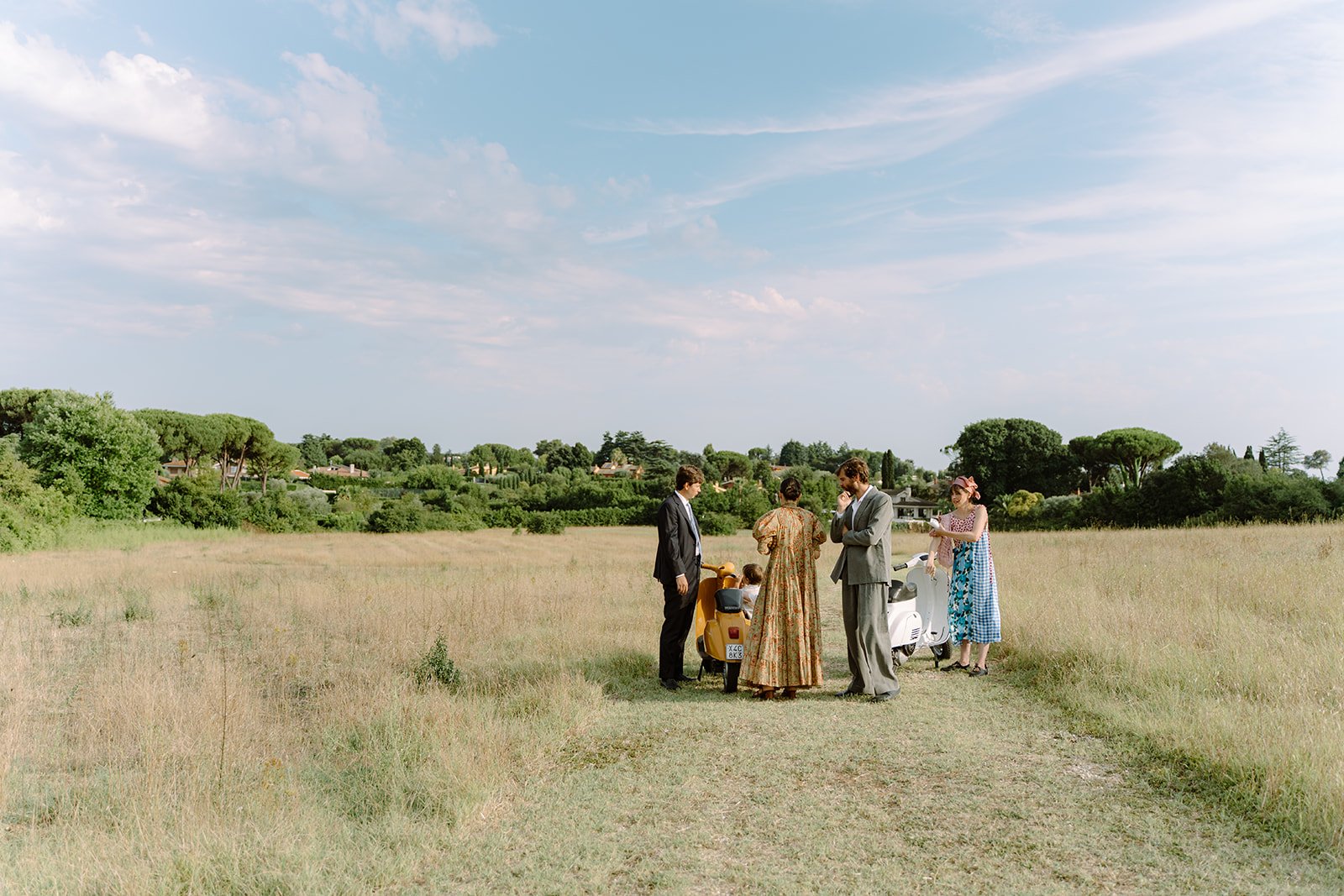 People dressed in vintage clothing standing and talking in a grassy field under a partly cloudy sky, with trees and houses in the distance.