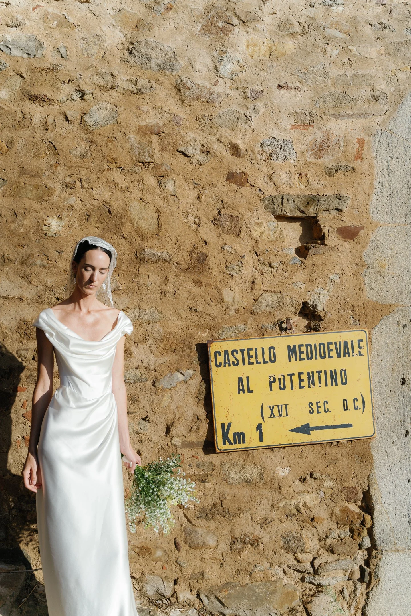 A woman in a white dress holding a bouquet of small white flowers, standing beside a yellow directional road sign against a stone wall.