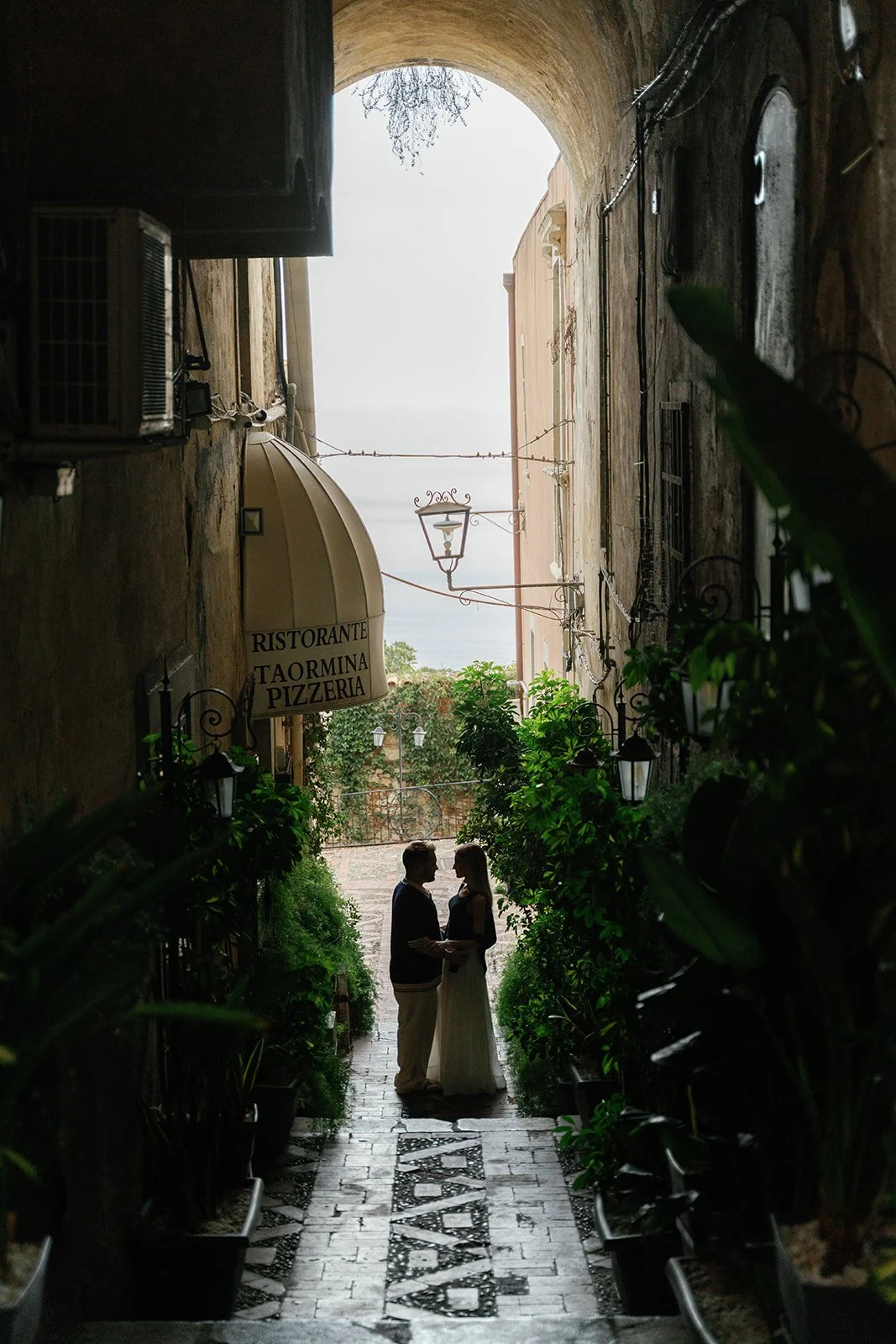 Silhouette of a couple holding hands in a narrow alley with plants and a sign for a restaurant and pizzeria, leading to an outdoor view of the sea.