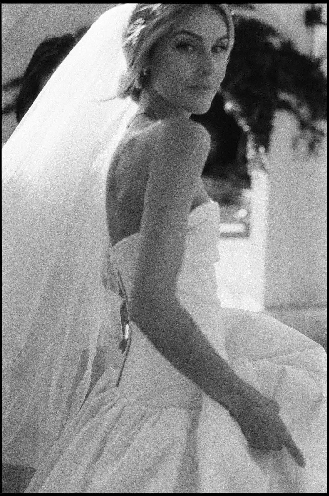 Black and white photo of a woman in a wedding dress with a veil, sitting and looking over her shoulder.