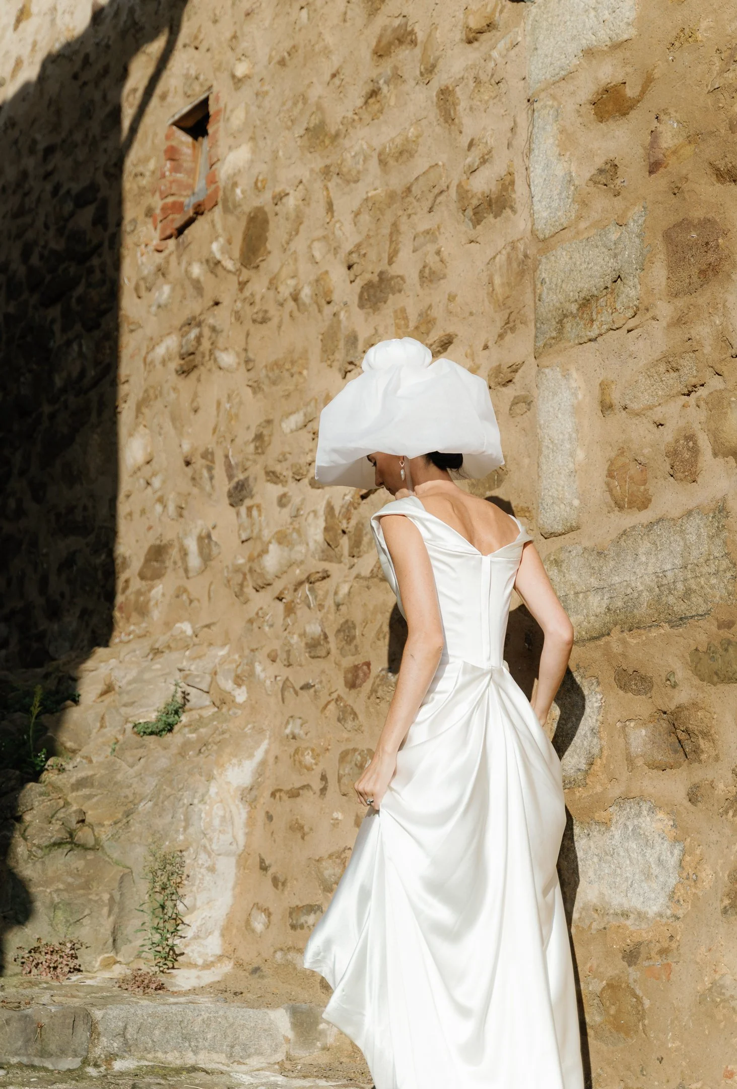 A woman in a white dress and large white hat stands near a stone wall, looking downward.
