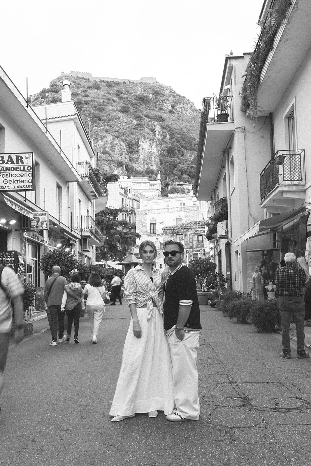 A black and white photo of a street scene with two people in vintage-style clothing centered in the foreground. A woman in a long skirt and striped blouse stands next to a man in white pants and a dark shirt with sunglasses. Several people walk on th