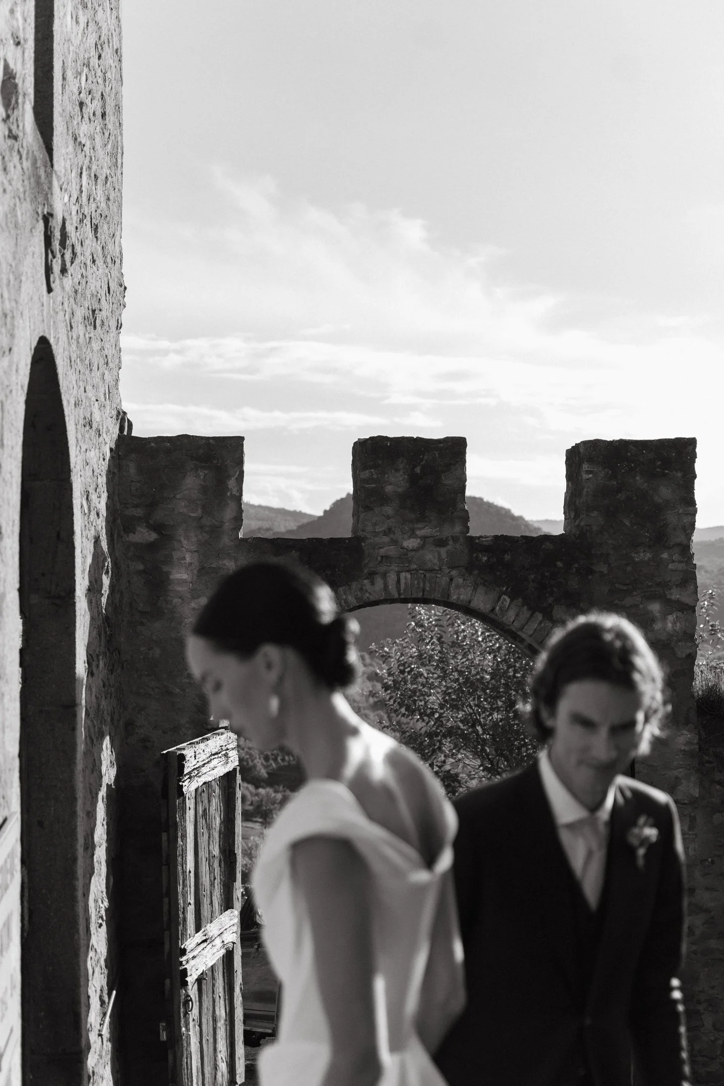Black and white photo of a woman and a man in formal attire walking past a stone building with a castle wall in the background.