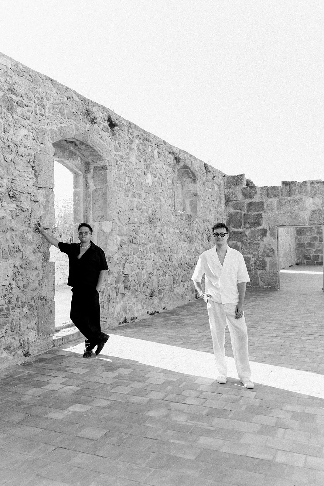 Two men standing in front of a stone wall with one wearing black and the other wearing white, both with short hair and glasses, in an outdoor setting with brick flooring.