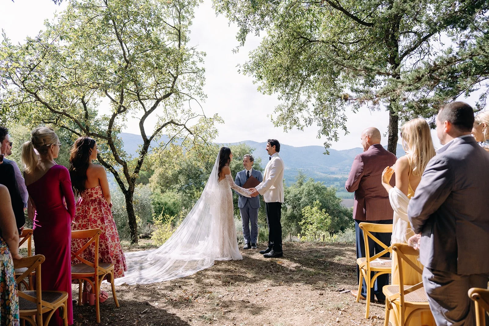 A wedding ceremony outdoors with a bride and groom holding hands, exchanging vows, surrounded by seated guests in a natural setting with trees and mountains in the background.