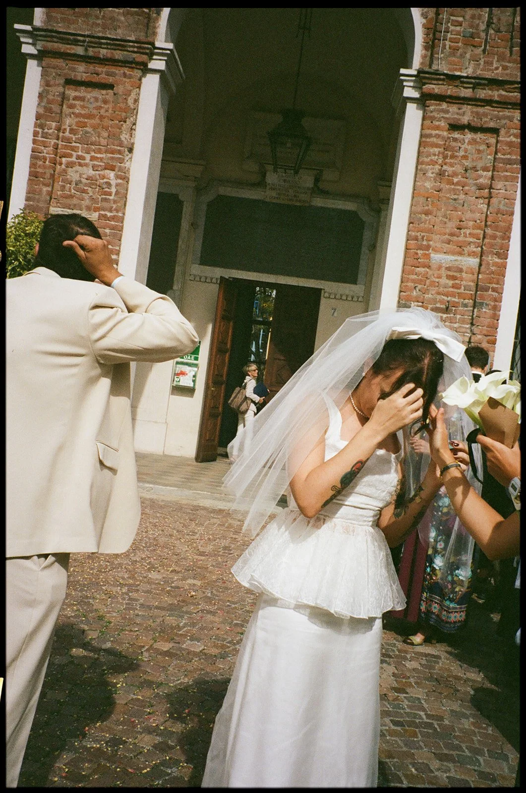 A woman in a white wedding dress with tattoos on her arms and a veil is holding her head while someone offers her white flowers during a wedding ceremony outside a brick building.