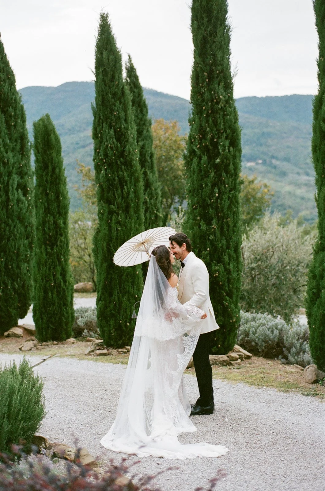 A bride and groom on their wedding day, standing outdoors on a gravel path surrounded by tall green cypress trees and mountains in the background. The bride wears a white lace wedding dress and veil, holding a white parasol. The groom wears a white tuxedo jacket and black pants, smiling at the bride.