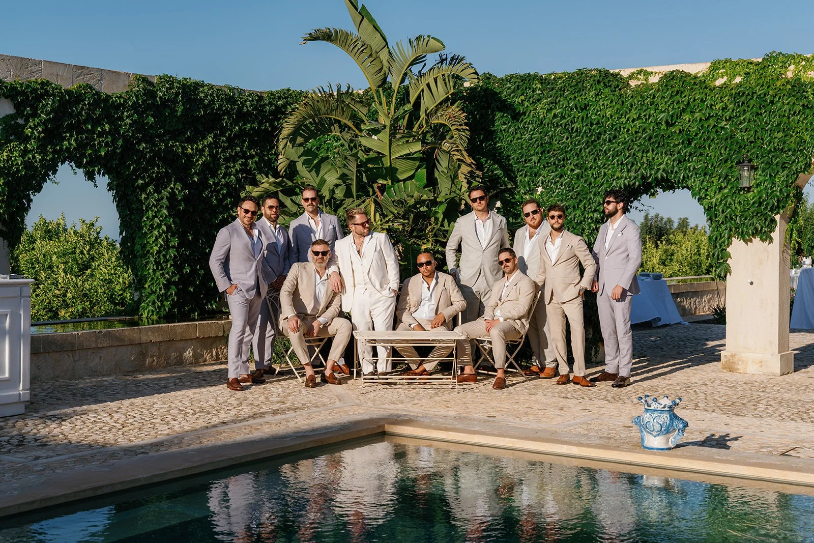 A group of men dressed in suits, posing by a poolside with lush green plants and a blue sky in the background.