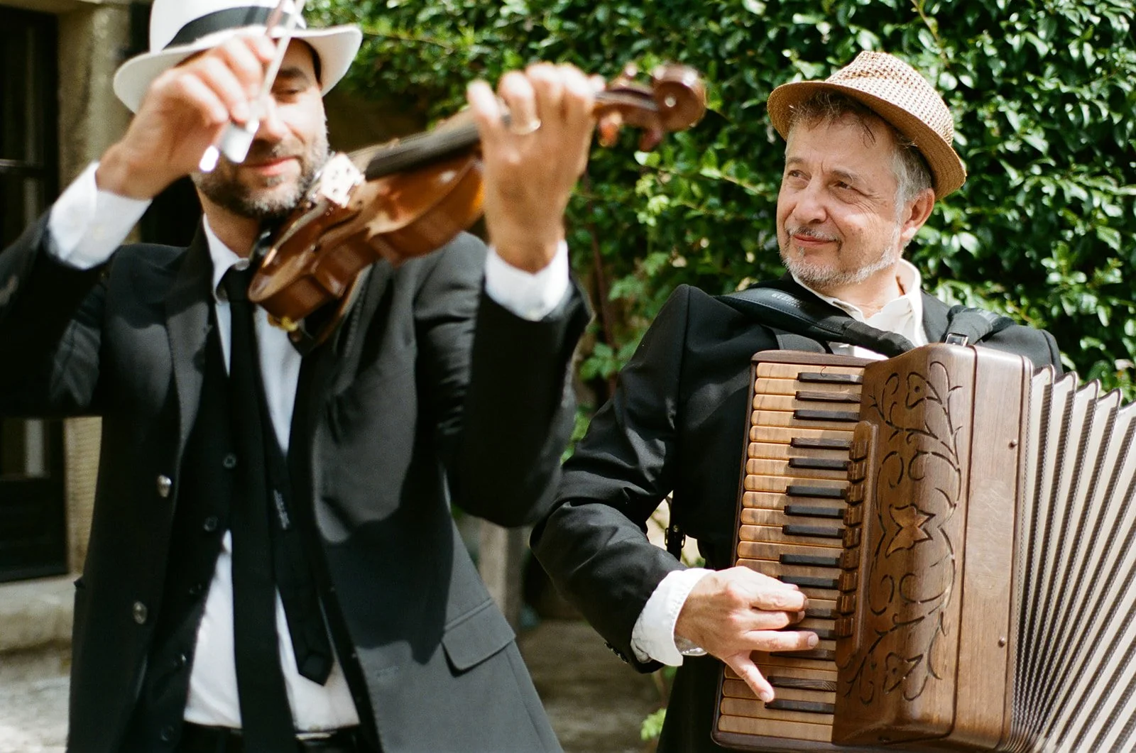 Two men dressed in dark suits and hats playing musical instruments outdoors with green foliage in the background. One man is playing a violin, and the other is playing an accordion.