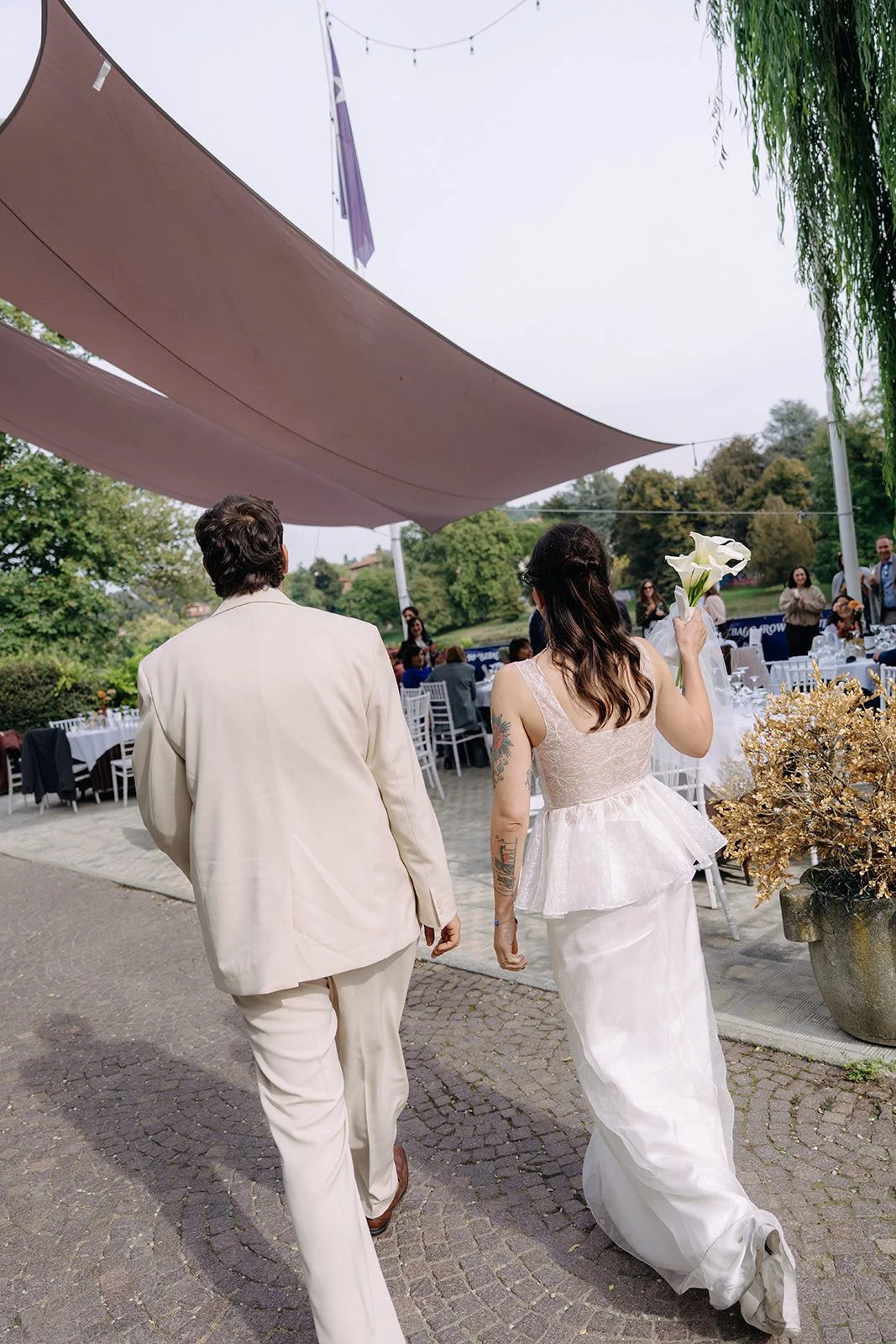 Bride in a white wedding dress holding calla lilies, walking with a person in a cream-colored suit at an outdoor wedding reception with tables and chairs under large fabric shades and string lights.