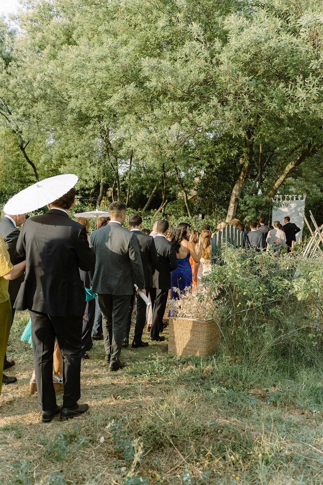People dressed in formal attire standing in a queue outdoors, under trees, waiting for an event to start.