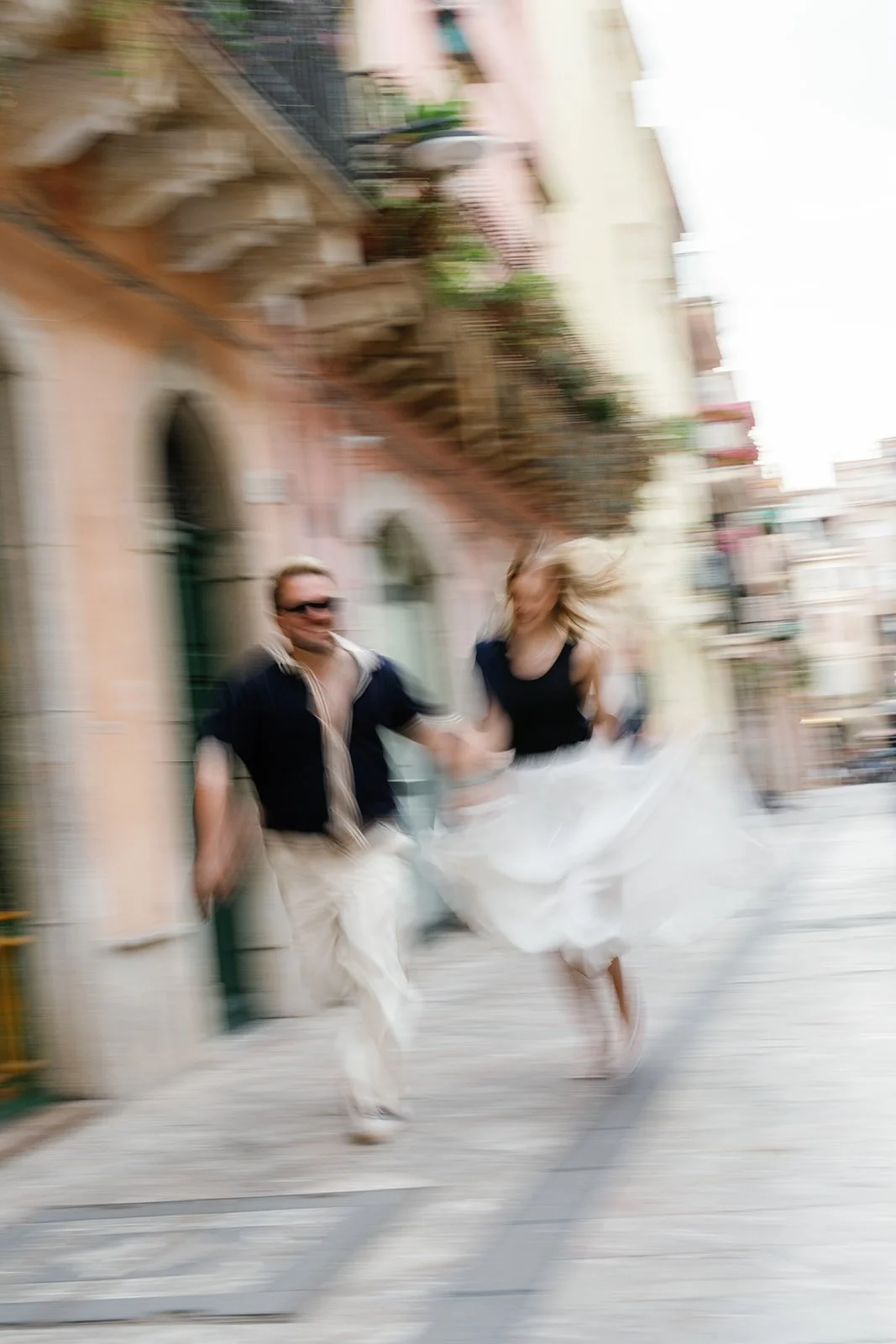 A blurry photo of a man and woman holding hands and running down a city street, with the woman in a white dress and the man in a dark shirt and light-colored pants.