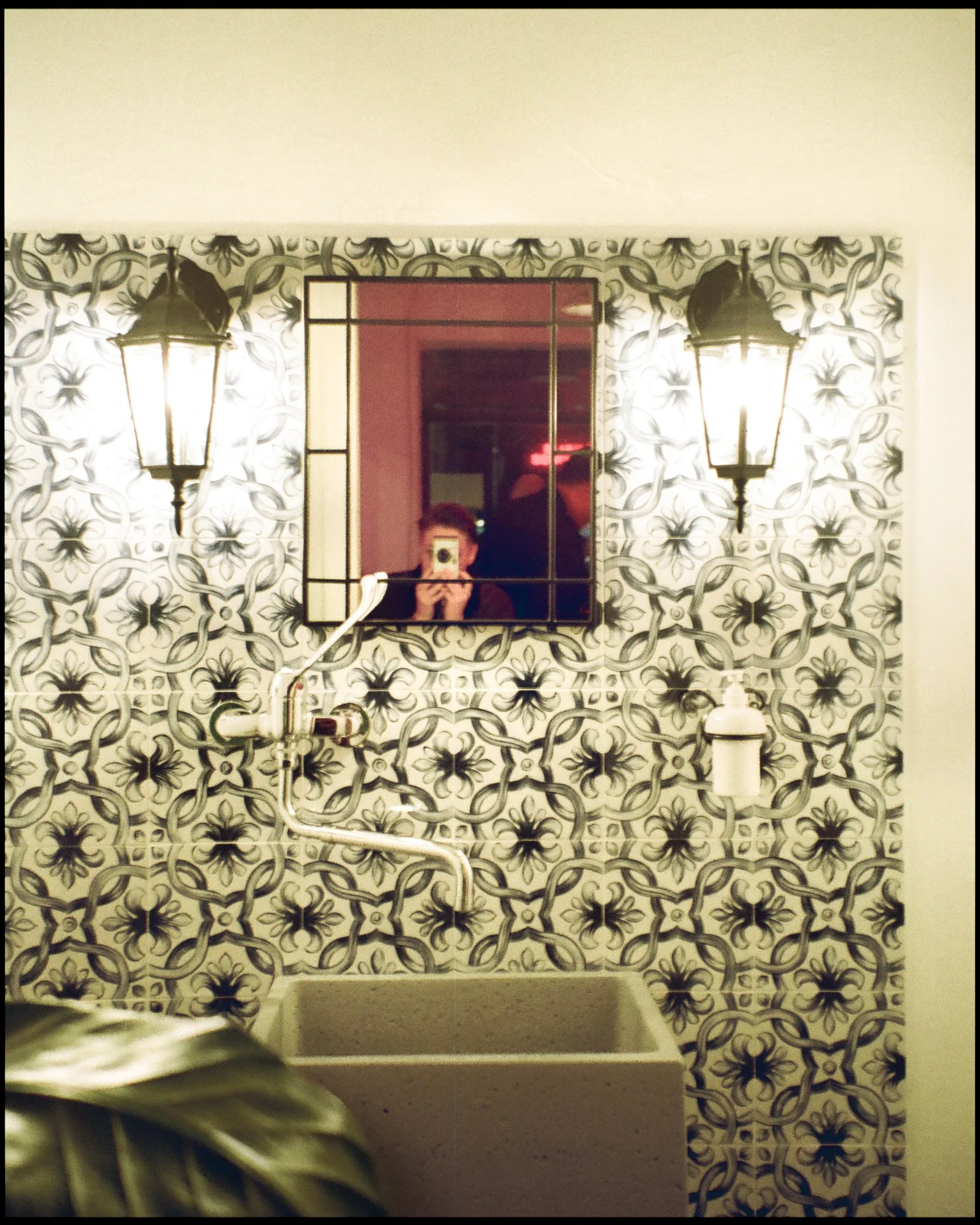 A wall with black and white patterned tiles, a mirror, two wall-mounted lanterns, and a soap dispenser. The mirror reflects a person taking a photo with a camera.