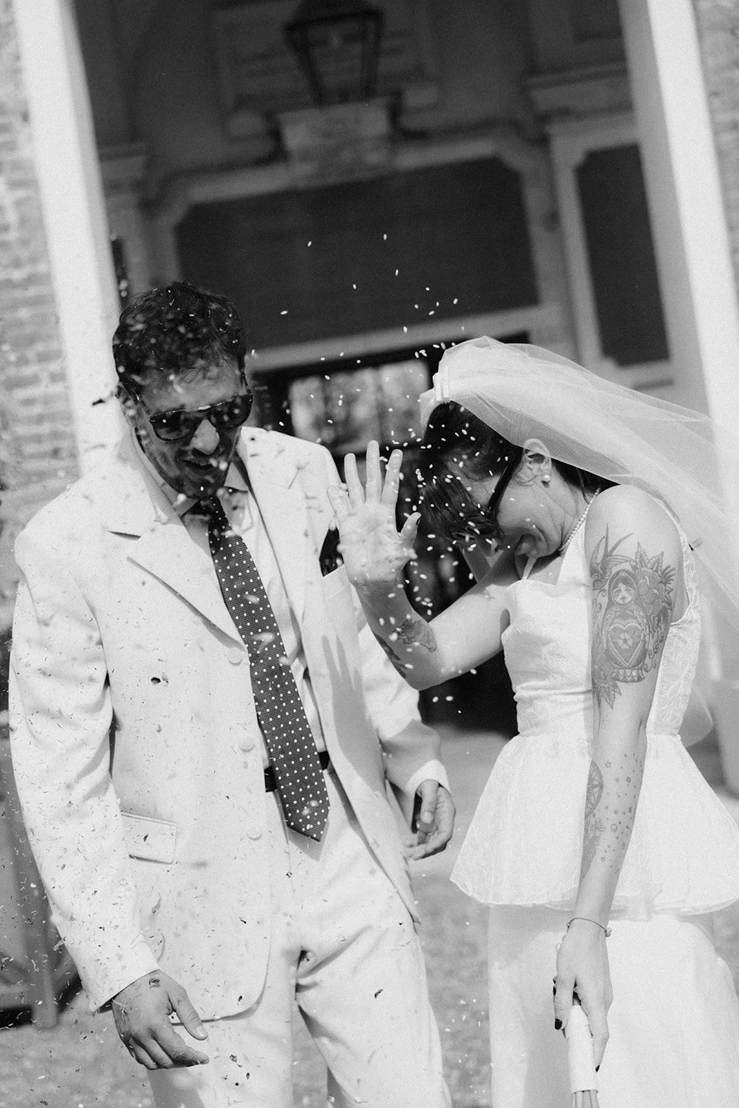 A bride and a man, both wearing sunglasses, share a joyful moment as confetti is thrown on them during a wedding celebration.